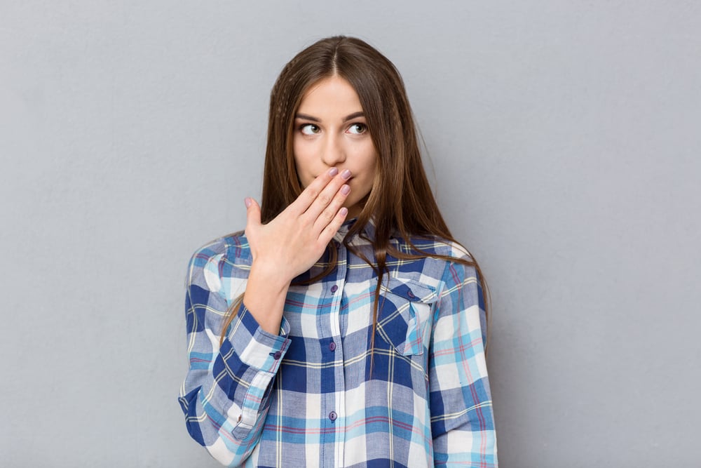 Portrait of a young woman covering her mouth with palm and looking away over gray background